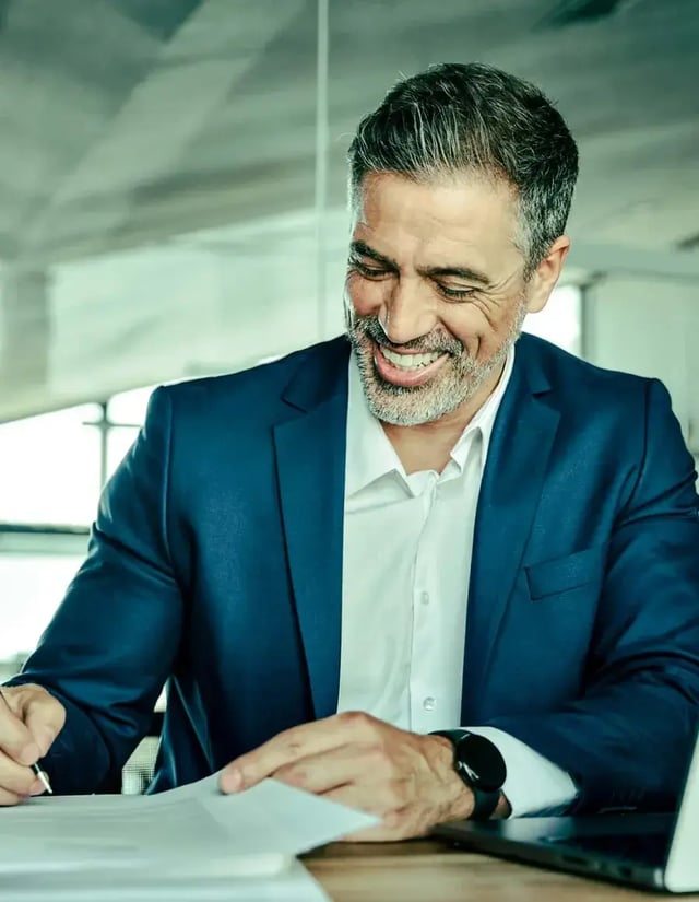 A smiling man in a suit jacket signs paperwork